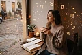 Susimąsčiusi moteris jaukiame megztinyje sėdi kavinėje prie lango, laikydama puodelį, o ore aplink ją sklando magiški, švelniai šviečiantys astrologiniai Zodiako ženklai.