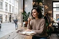 Savimi pasitikinti ir rami moteris sėdi kavinėje prie lango, geria kavą ir skaito knygą, o aplink ją ore sklando šviečiantys auksiniai astrologiniai Zodiako ženklai.