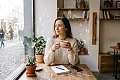 Elegantiška moteris geria kavą šviesioje aplinkoje, o aplink ją ore švelniai švyti sėkmę nešantys skaičiai ir Zodiako simboliai.