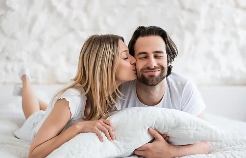 Miegamajame besišypsanti pora – moteris bučiuoja vyrą į žandą. Iliustracija straipsniui apie intymų suderinamumą pagal Zodiako ženklus