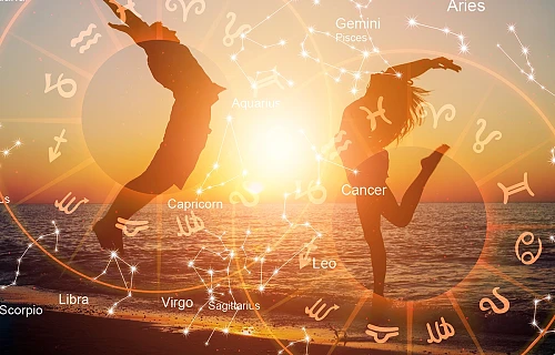Moteris ir vyras šoka į viršų jūros pakrantėje, aplink juos pavaizduoti du zodiako ženklų horoskopo ratai.